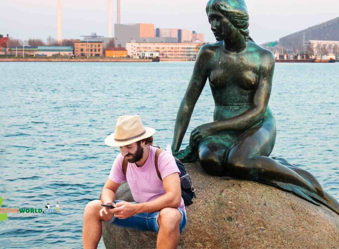 A serene scene capturing a Lestacworld tourist seated by the iconic Little Mermaid statue in Copenhagen. The tourist is peacefully gazing at the sculpture, which rests on a rock by the waterside. The statue, a symbol of Copenhagen, depicts a mermaid looking out to sea, surrounded by a calm and picturesque harbor.