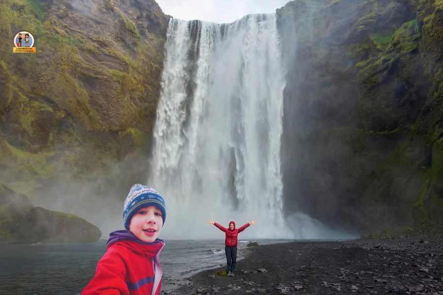 Child taking a selfie with Skógafoss Waterfall in the background, while her mother approaches from a distance, Iceland South Coast one-day tour from Reykjavik.